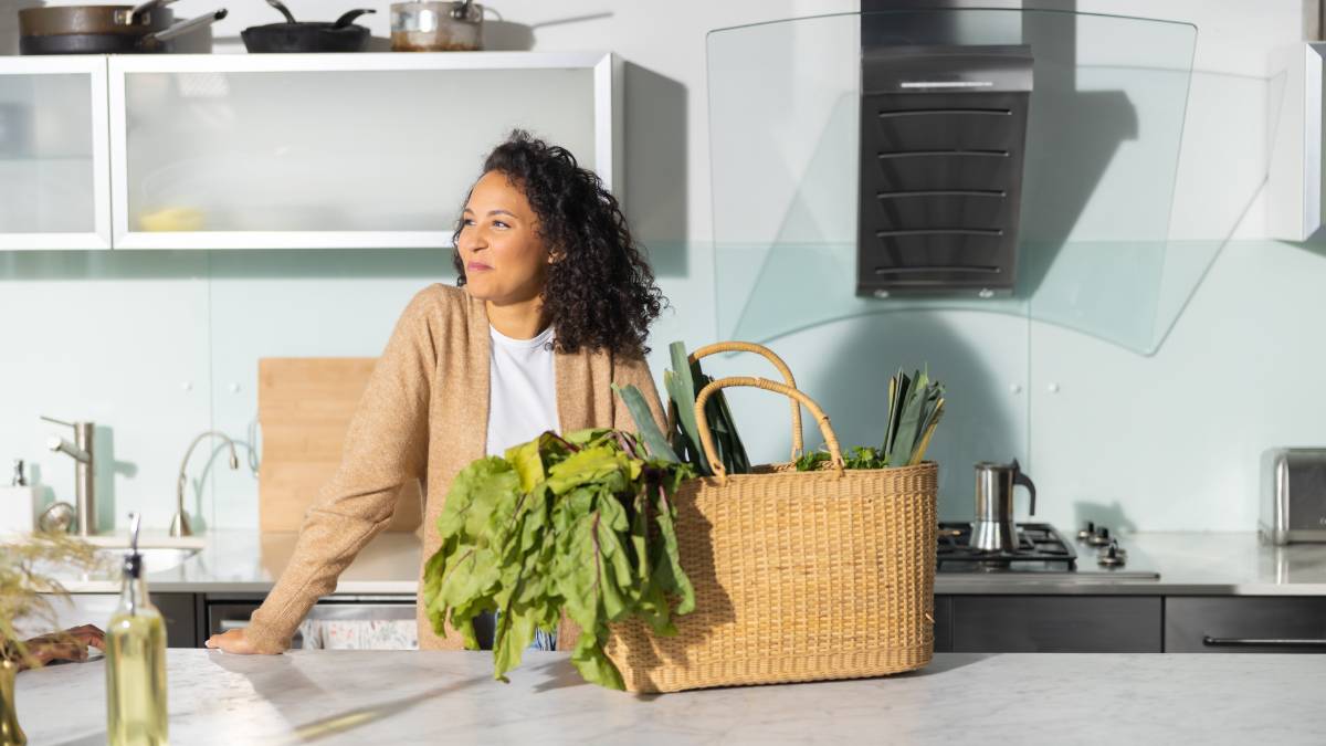 Woman in her kitchen smiling with healthy vegetables in a woven basket