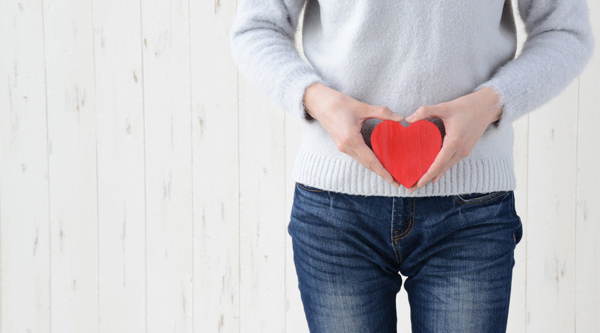 Person holding a red heart shape with hands over their gut against a white wooden background.
