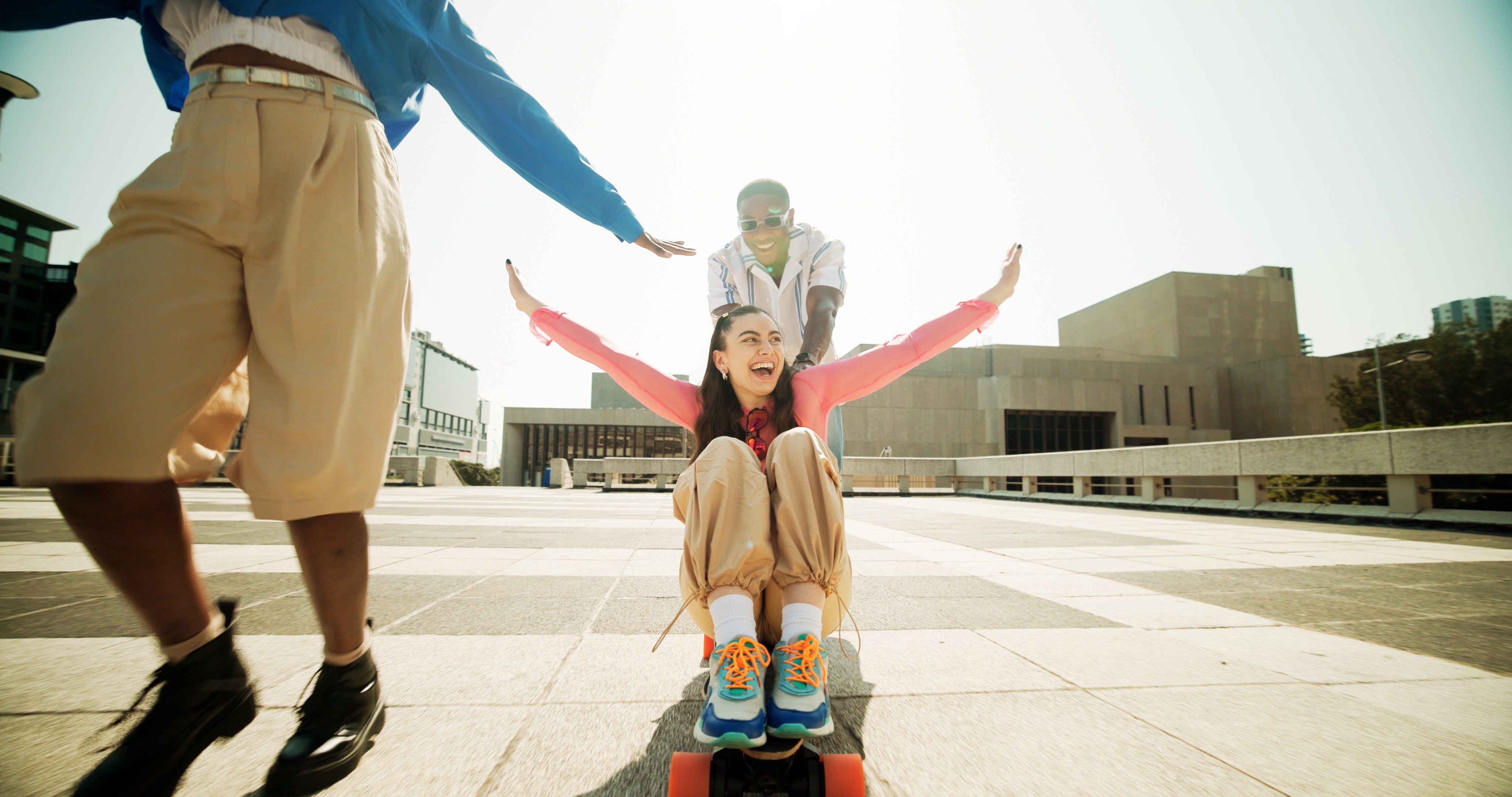 A lifestyle scene outdoors showing a young woman in a pink jacket and beige pants riding a skateboard, with a young man behind her and another person in the foreground, set against a bright, sunny urban plaza with modern buildings.