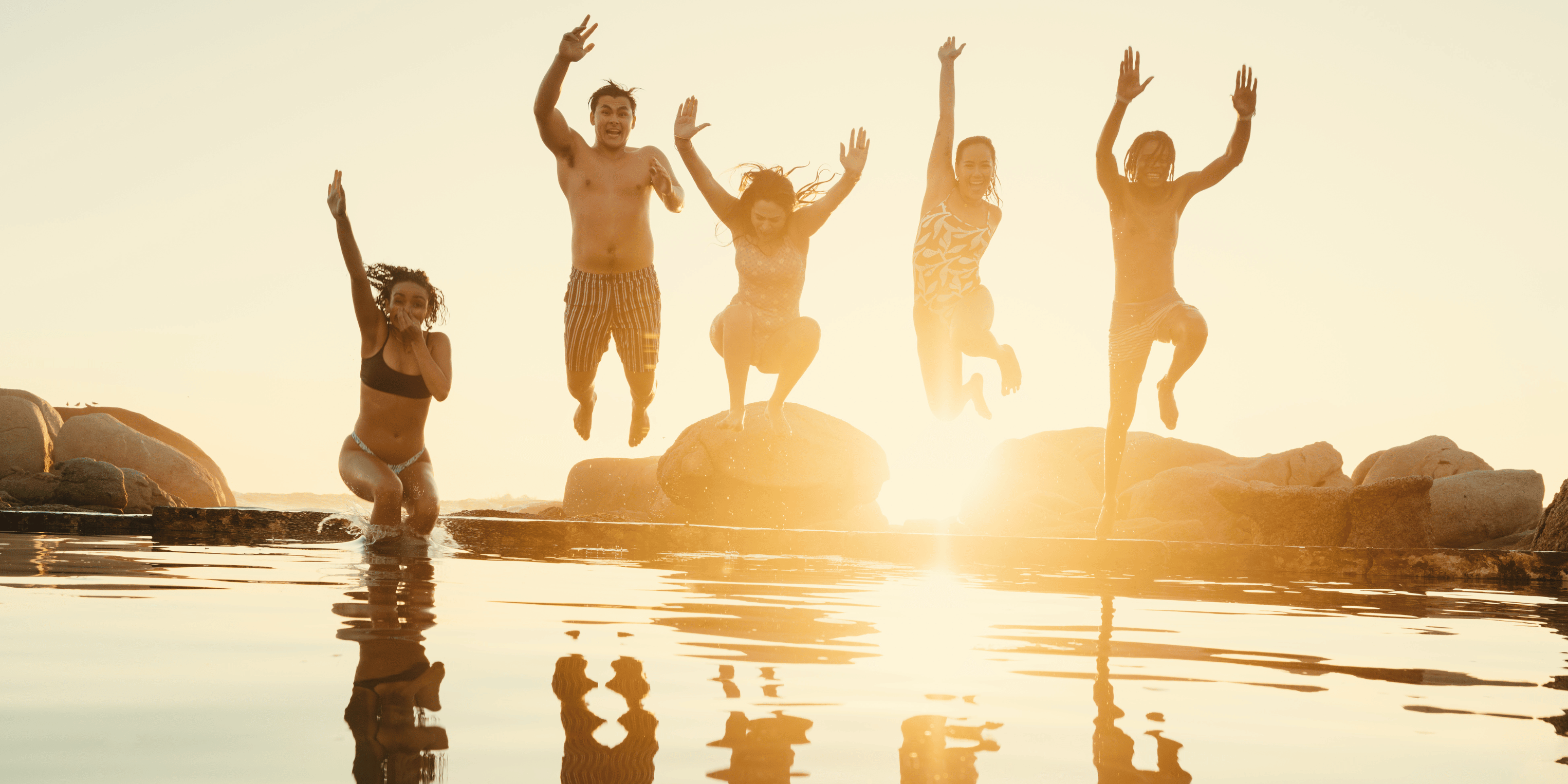 A group of five people jumping joyfully into shallow water at sunset, with their reflections clearly visible on the calm surface. The scene is backlit by a bright golden sun, creating silhouettes against a warm sky.
